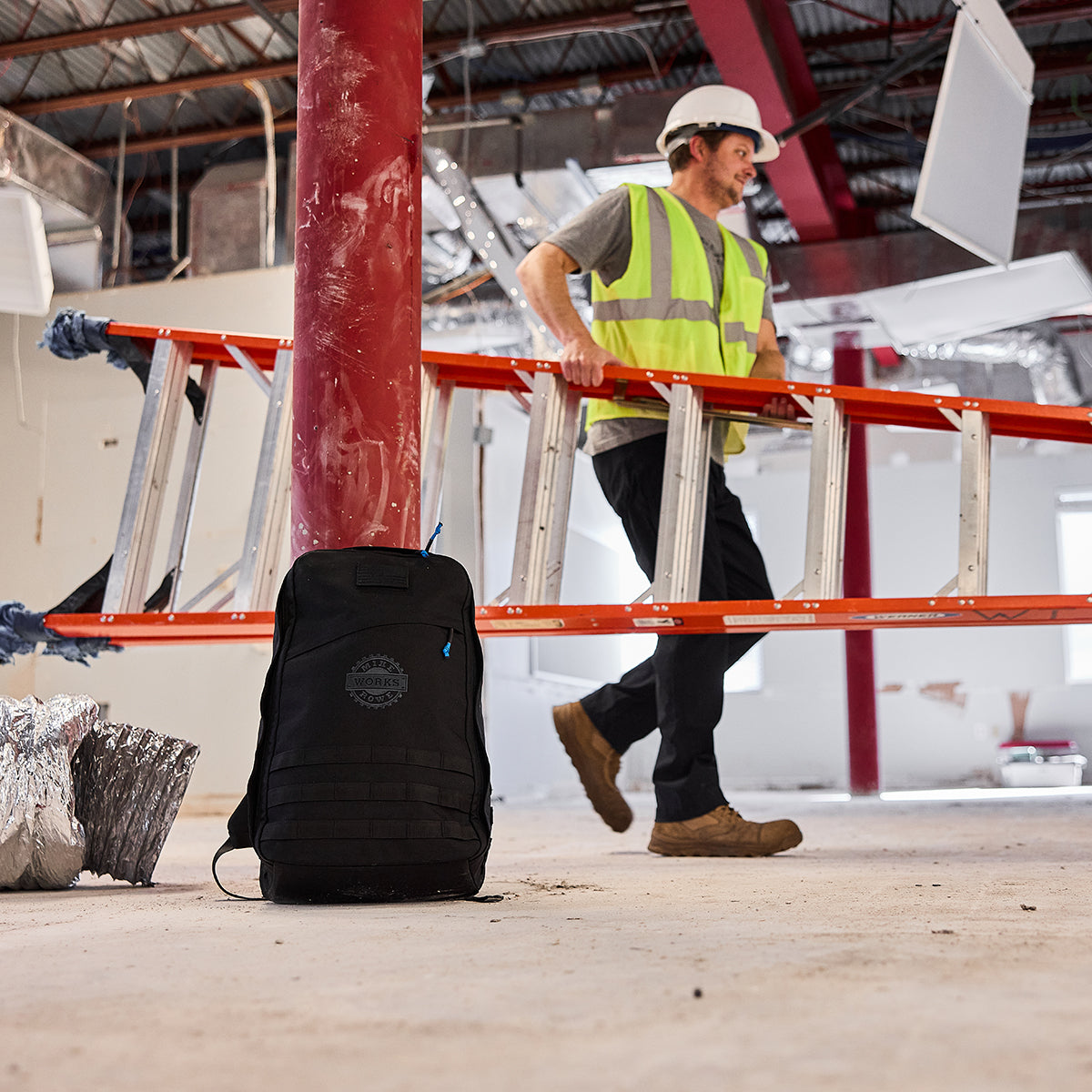 A construction worker in a hard hat and vest carries a ladder through an industrial site, showcasing American craftsmanship; the limited-edition GR1 USA - GORUCK X mikeroweWORKS backpack is slung in front, embodying the foundation's spirit.