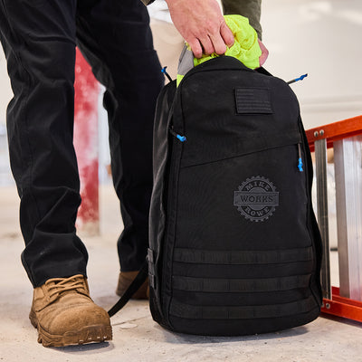 Near a ladder, someone places a high-visibility yellow jacket into a GR1 USA GORUCK X mikeroweWORKS backpack, known for its American craftsmanship and the values of the mikeroweWORKS Foundation.