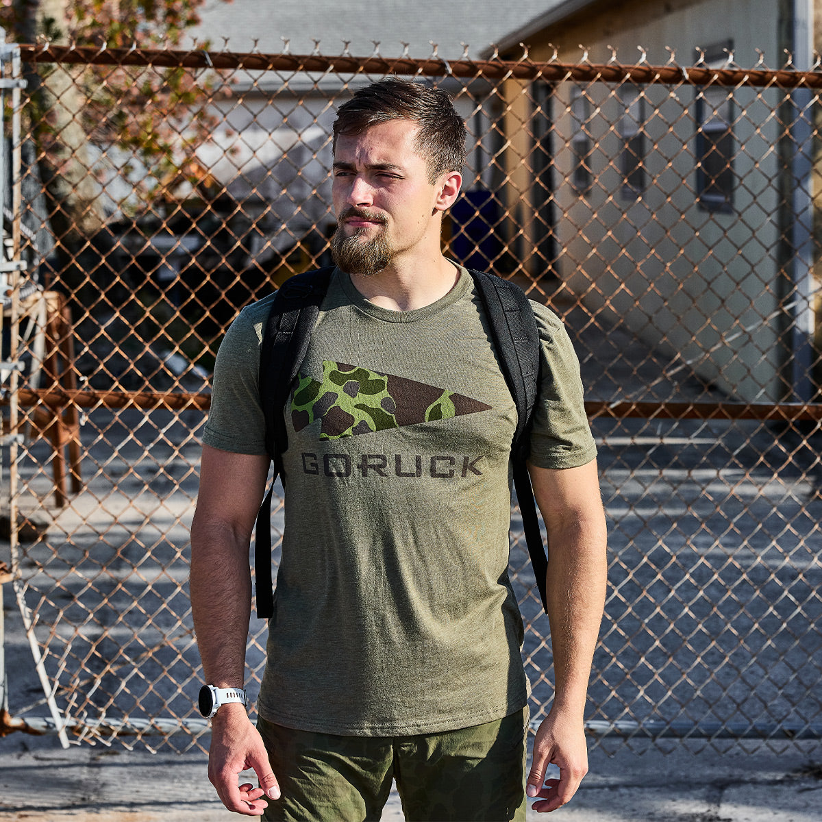 A man with a beard stands in front of a chain-link fence, wearing the olive green GORUCK Spearhead Tee - Tri-Blend by vendor-unknown, which boldly features the iconic spearhead logo reminiscent of its Special Forces heritage. He carries a backpack over his shoulder against the backdrop of a sunlit building.