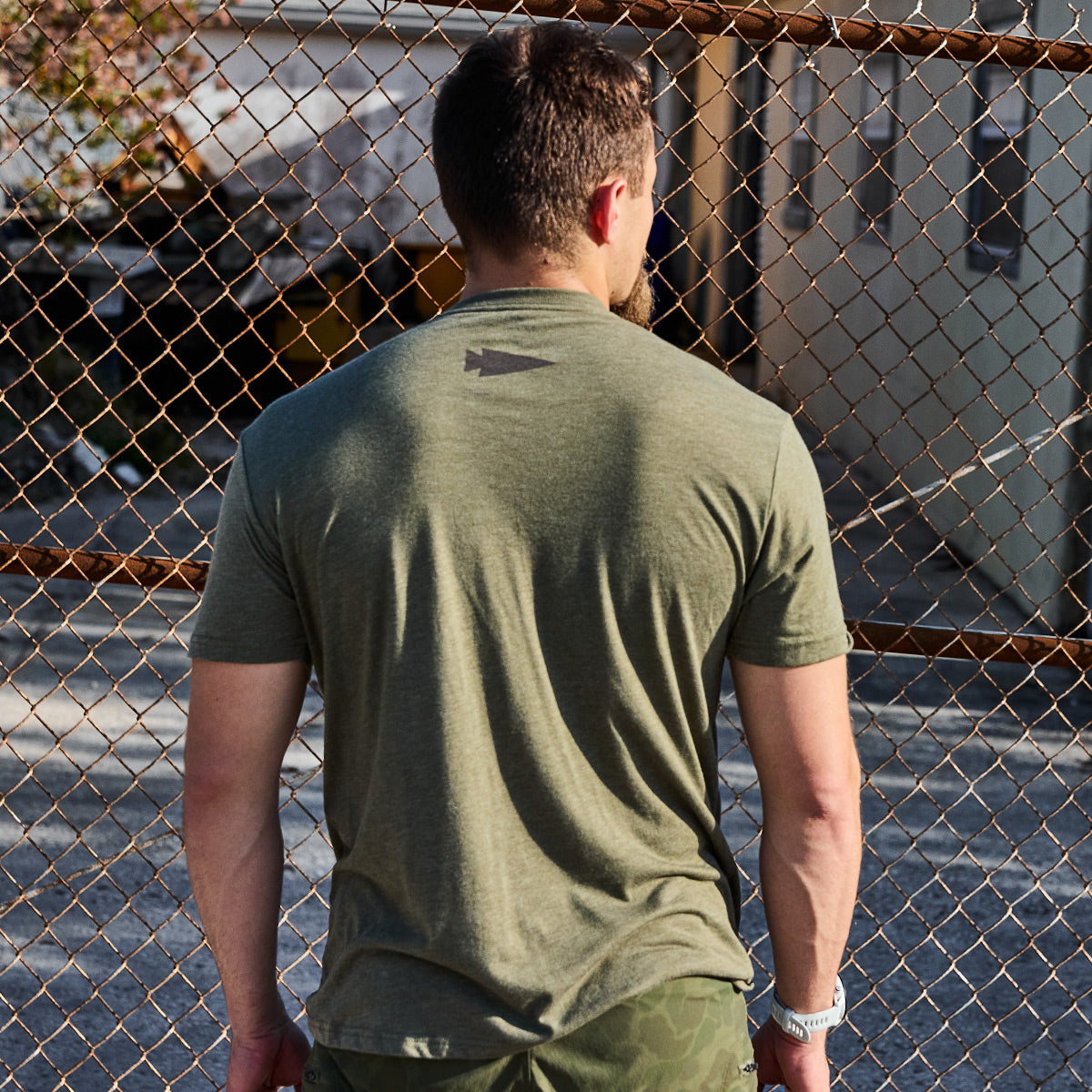 A man with Special Forces roots stands with his back to the camera wearing a GORUCK Spearhead Tee - Tri-Blend in olive green and camo shorts. He is positioned in front of a chain-link fence, where sunlight casts shadows on the ground, all while a blurred building in the background captures the rugged spirit of vendor-unknown.