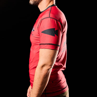 A man wearing the Murder Yoga Rash Guard by GORUCK, featuring a red design with a black arrow pattern and engineered for moisture-wicking comfort, stands against a black background.