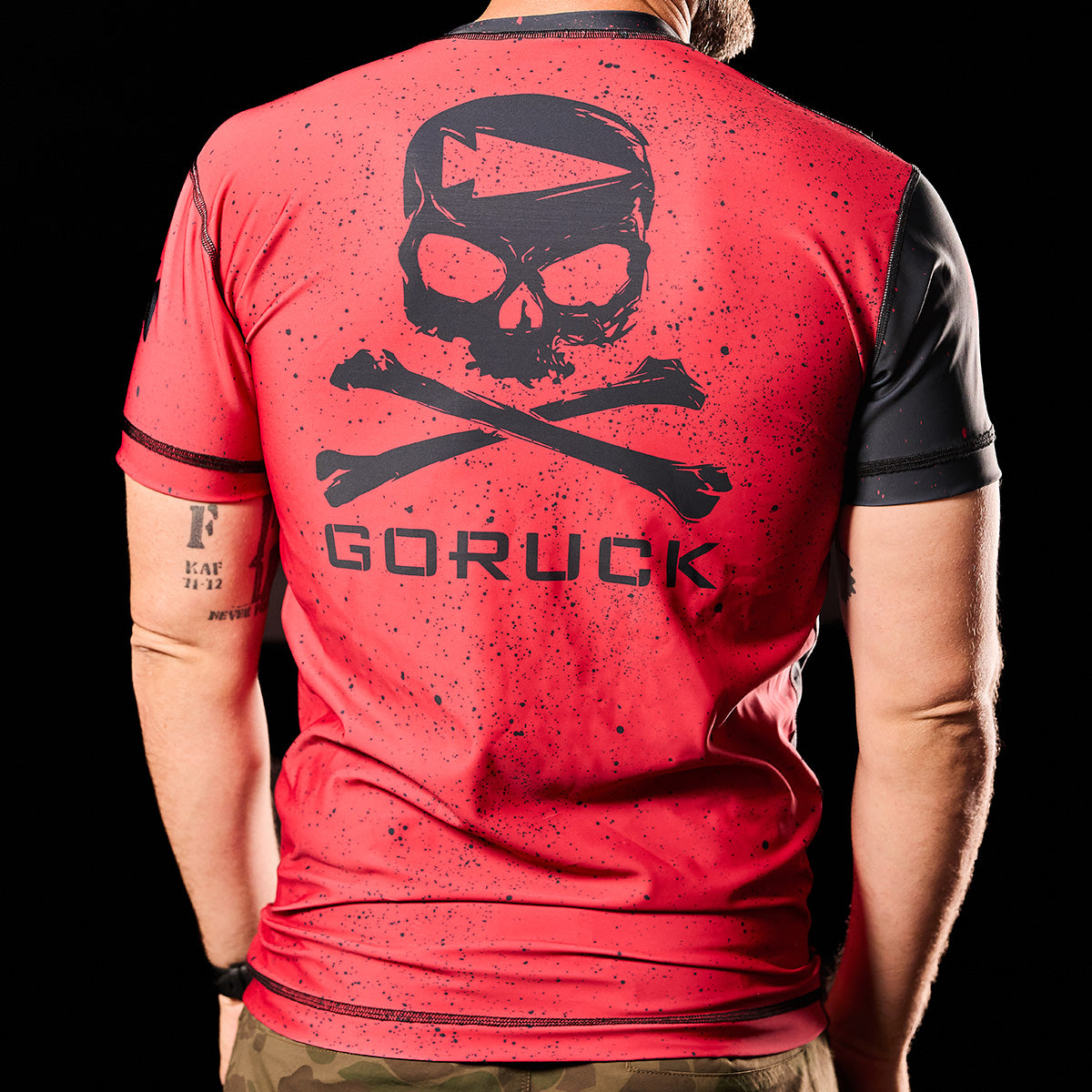 A person wears a Murder Yoga Rash Guard - Built By SCARS from GORUCK, showcasing a red t-shirt with a skull and crossbones design and "GORUCK" text on the back, crafted from moisture-wicking WARPWICK textile for ultimate comfort.