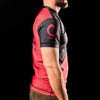 A man stands against a black background wearing the Murder Yoga Rash Guard - Built By SCARS from GORUCK, featuring red and black athletic design with "ORIGIN" on the sleeve for moisture-wicking comfort.