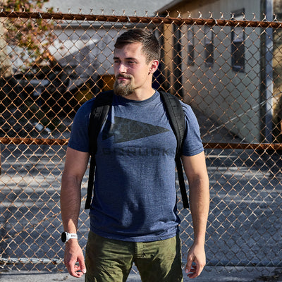 A person wearing a GORUCK Spearhead Tee in blue and green pants stands outside, carrying a black backpack featuring the GORUCK spearhead. They have short hair and a beard and are wearing a watch. A chain-link fence and a building are in the background.