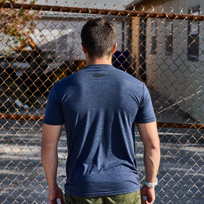 A man wearing the GORUCK Spearhead Tee - Tri-Blend from vendor-unknown stands with his back to the camera in front of a chain-link fence, exuding ruggedness. A building and trees form the backdrop, hinting at his Special Forces roots, as he pairs the premium blue T-shirt with green pants.