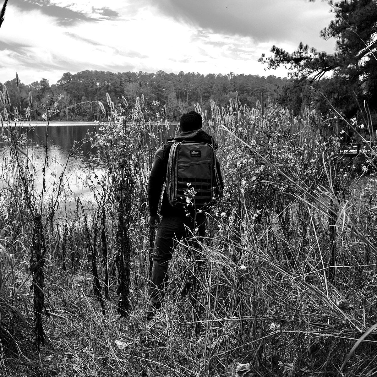 A person equipped with the GR2 - Dyneema by GORUCK stands on a grassy lakeside, looking at the water. The scene is enveloped by tall grasses and trees under a cloudy sky. The backpack, crafted from abrasion-resistant Dyneema®, complements the serene and contemplative mood of the black-and-white image perfectly.