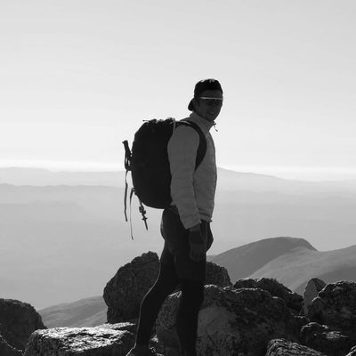 A black and white photo captures the silhouette of a person with a GR3 - Dyneema carry-on backpack by GORUCK, standing atop a rocky mountain peak. The clear sky accentuates the vast landscape and distant hills, while highlighting the textures of the rocks and Dyneema material.