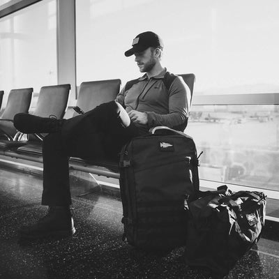 A person dressed in dark clothing and a baseball cap sits in an airport terminal, checking their phone. Beside them are two large bags, including a GORUCK Kit Bag - Dyneema. The scene is captured in striking black and white, evoking the stealth of Special Forces in transit.