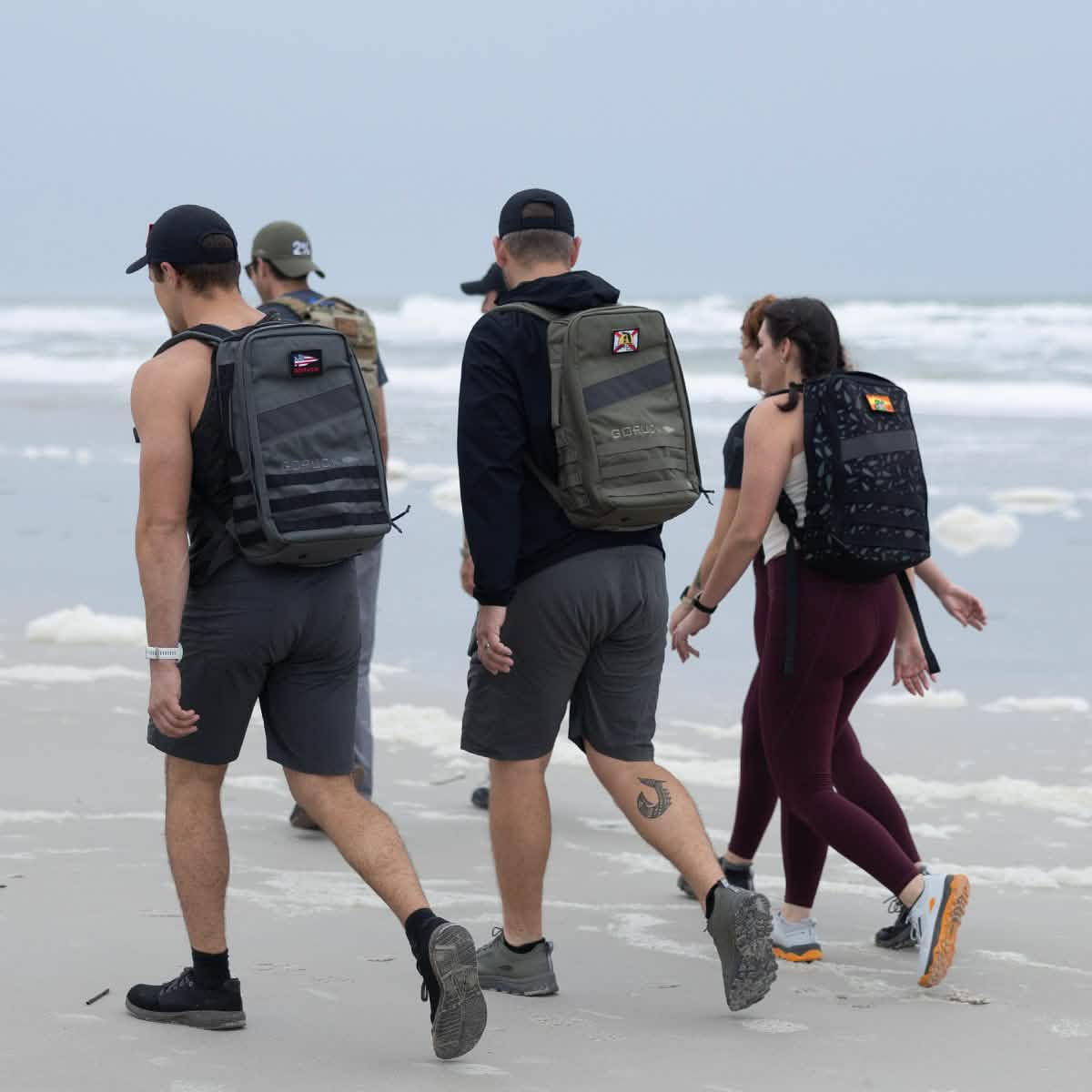 A group of people, sporting backpacks and cozy GORUCK Merino Challenge Socks - Ankle, stroll along a sandy beach. The sky is overcast, with ocean waves rolling in the background. Dressed in casual outdoor attire like shorts and leggings, they enjoy their adventure to its fullest.