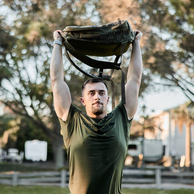 Wearing the GORUCK Men’s Performance Tee - ToughMesh in green, a man is outdoors, hoisting a green backpack over his head. He stands on grass with trees and a fence behind him. The clear sky indicates it's a sunny day, ideal for putting the ToughMesh™ fabric to the test.