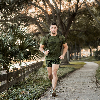 A man in athletic gear wearing the GORUCK Men’s Performance Tee - ToughMesh jogs along a paved path lined with trees and a wooden fence. The sun filters through the branches, creating a warm atmosphere. He appears focused as he moves toward the camera.