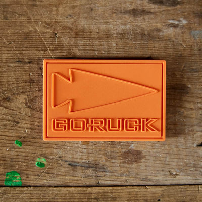 A PVC Patch - GORUCK, designed in a vibrant orange rectangle and made from sturdy plastic, features an embossed spearhead motif with the word GORUCK, all placed against a wooden backdrop.