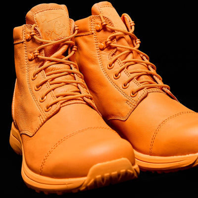 A pair of bright orange MACV-1 Traveler x Carryology - Berserker Viking boots by GORUCK, designed for rugged terrain with thick soles, making them perfect for travel and adventure. Set against a black background, these water-resistant leather boots showcase visible stitching and embossed branding on the tongue.