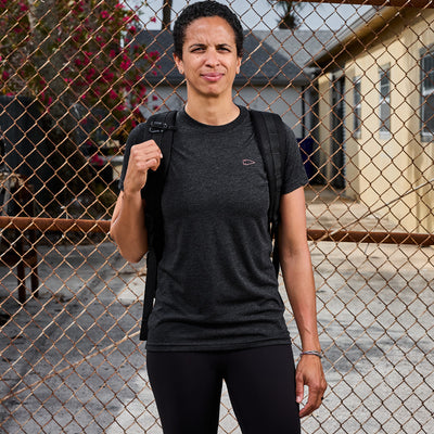 A person with curly hair stands outside a chain-link fence, wearing a dark Subtle Spearhead Tee - Tri-Blend and a backpack. They gaze ahead with a slight smile, while buildings and foliage form the backdrop.