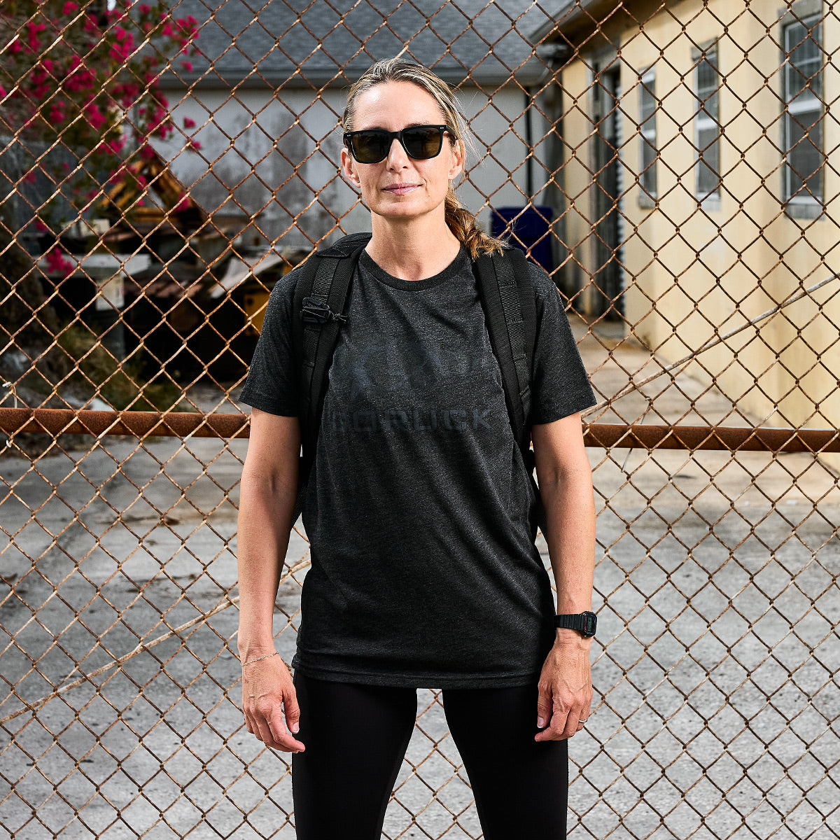 A person wearing GORUCK Spearhead sunglasses, a black GORUCK Spearhead Tee—Tri-Blend from vendor-unknown, and a backpack stands in front of a chain-link fence. Premium tri-blend houses and a small tree frame the background on a cloudy day.