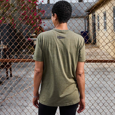 A person with short curly hair stands with their back to the camera, wearing an olive green GORUCK Spearhead Tee - Tri-Blend and black pants. They face a chain-link fence, while buildings and a bush adorned with red flowers enhance the backdrop.