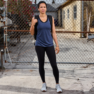 A person stands outdoors on a paved surface, wearing a blue Women's Racerback Tank - Tri-Blend by GORUCK, black leggings, and gray sneakers. With a backpack slung over one shoulder, they seem ready for a workout. Behind them, a chain-link fence and buildings loom under an overcast sky.