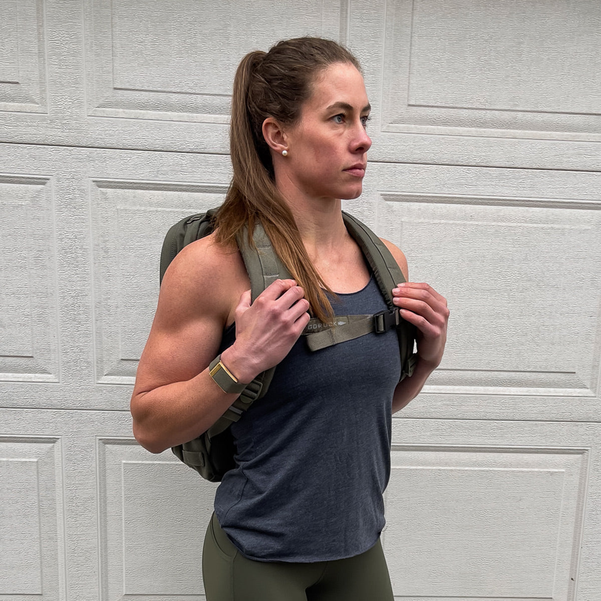 A woman with long hair in a ponytail stands against a light gray paneled wall, wearing a GORUCK Women's Racerback Tank - Tri-Blend and leggings, paired with a green backpack. She gazes ahead, embodying the spirit of determination.