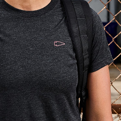 A person is wearing the Subtle Spearhead Tee - Tri-Blend from vendor-unknown, with its dark gray fabric showcasing a discreet spearhead logo on the chest. Complementing this look is a black backpack strap resting over the shoulder, all set against a chain-link fence backdrop that echoes Special Forces training scenes.