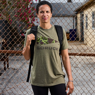 Standing confidently in front of a chain-link fence, the person wears an olive green GORUCK Spearhead Tee - Tri-Blend, paired with a black backpack. They are outdoors on a sunny day with buildings in the background, capturing the rugged spirit inspired by Special Forces roots and offered by vendor-unknown.