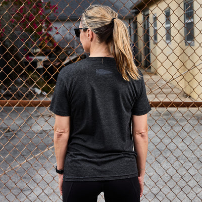 A woman with a ponytail stands facing a chain-link fence, wearing the GORUCK Spearhead Tee - Tri-Blend in black and sunglasses. The background features a building and paved area, evoking her Special Forces roots with the GORUCK spearhead vibe.