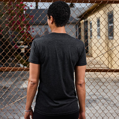 A person with short curly hair, wearing the Subtle Spearhead Tee - Tri-Blend in dark gray from vendor-unknown, stands with their back to the camera in front of a chain-link fence. Behind the fence are buildings and a partially cloudy sky, reminiscent of a scene straight from Special Forces training grounds.