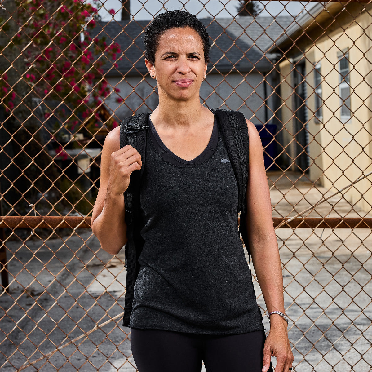 A person with short curly hair, wearing a Women's Racerback Tank - Tri-Blend from GORUCK and carrying a backpack, stands in front of a chain-link fence. They seem to be outdoors in a residential area with buildings behind them, likely preparing for a workout.