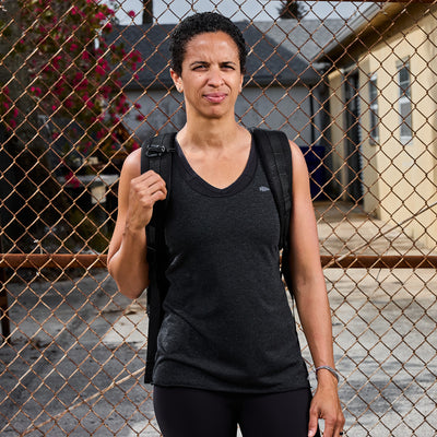 A person with short curly hair, wearing a Women's Racerback Tank - Tri-Blend from GORUCK and carrying a backpack, stands in front of a chain-link fence. They seem to be outdoors in a residential area with buildings behind them, likely preparing for a workout.