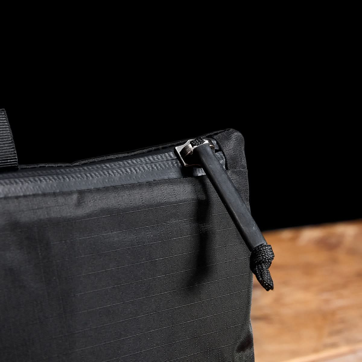 Close-up of a black textured Packable Bullet Ruck - Ripstop ROBIC® bag with a zipper and pull tab, against a black and wooden background.