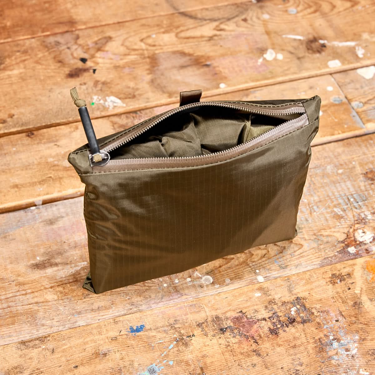 A Packable Bullet Ruck made of Ripstop ROBIC® nylon in green holds a pen, elegantly placed upright on a wooden surface with paint splatters.