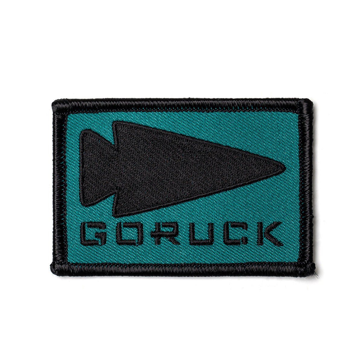 The GORUCK Spearhead Patch features a teal background with a prominent black arrowhead shape pointing to the right. Below this design, the GORUCK logo appears in bold black capital letters. The patch is bordered with a durable black edge and includes a convenient VELCRO backing for easy attachment.