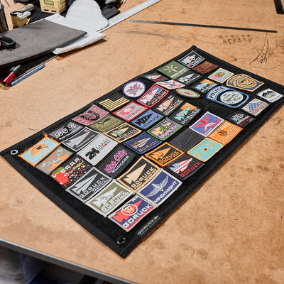 A GORUCK Spearhead Patch Board from GORUCK is displayed on a wooden table, featuring a collection of colorful embroidered patches. Some patches are affixed to the sturdy 1000D Cordura surface, while others adhere to the velcro panel, forming a dynamic tapestry of designs and textures.