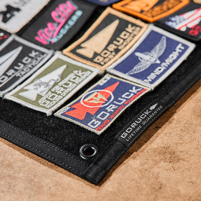 The GORUCK Spearhead Patch Board features a black 1000D Cordura fabric panel adorned with a variety of colorful patches, including one labeled "GORUCK," and comes equipped with a velcro panel.