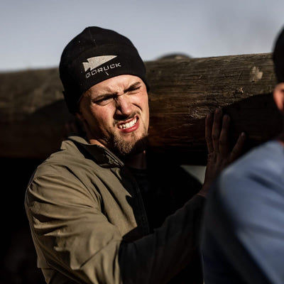 Wearing a Performance Beanie with the GORUCK Logo, a person in an olive jacket exerts effort as they carry a large wooden log on their shoulder, reminiscent of a GORUCK Event. The blurred background accentuates the subject's strained expression under the bright outdoor light.