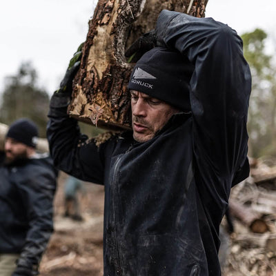 During a GORUCK event in the forest, an individual wearing a black jacket and the Performance Beanie - GORUCK Logo is seen carrying a large piece of wood on their shoulder, showcasing their cold weather rucking gear. Another person, similarly outfitted, stands in the background, embodying the spirit of cold weather rucking.