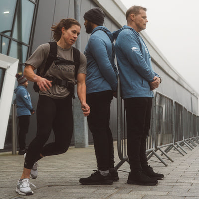 A woman in athletic gear speeds by flaunting Race Day Socks, known for maximum breathability, while two men in blue jackets stand beside a metal barricade.