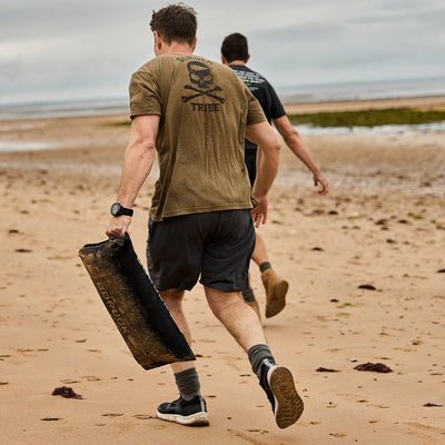 Two men are running on a sandy beach. The man in the foreground, wearing a green T-shirt with an emblem, carries a heavy black object. Under an overcast sky and amidst seaweed-strewn sand at low tide, they don the GORUCK Men's Rough Runner - Black + White lightweight running shoes, perfect for any Rough Runner challenge.