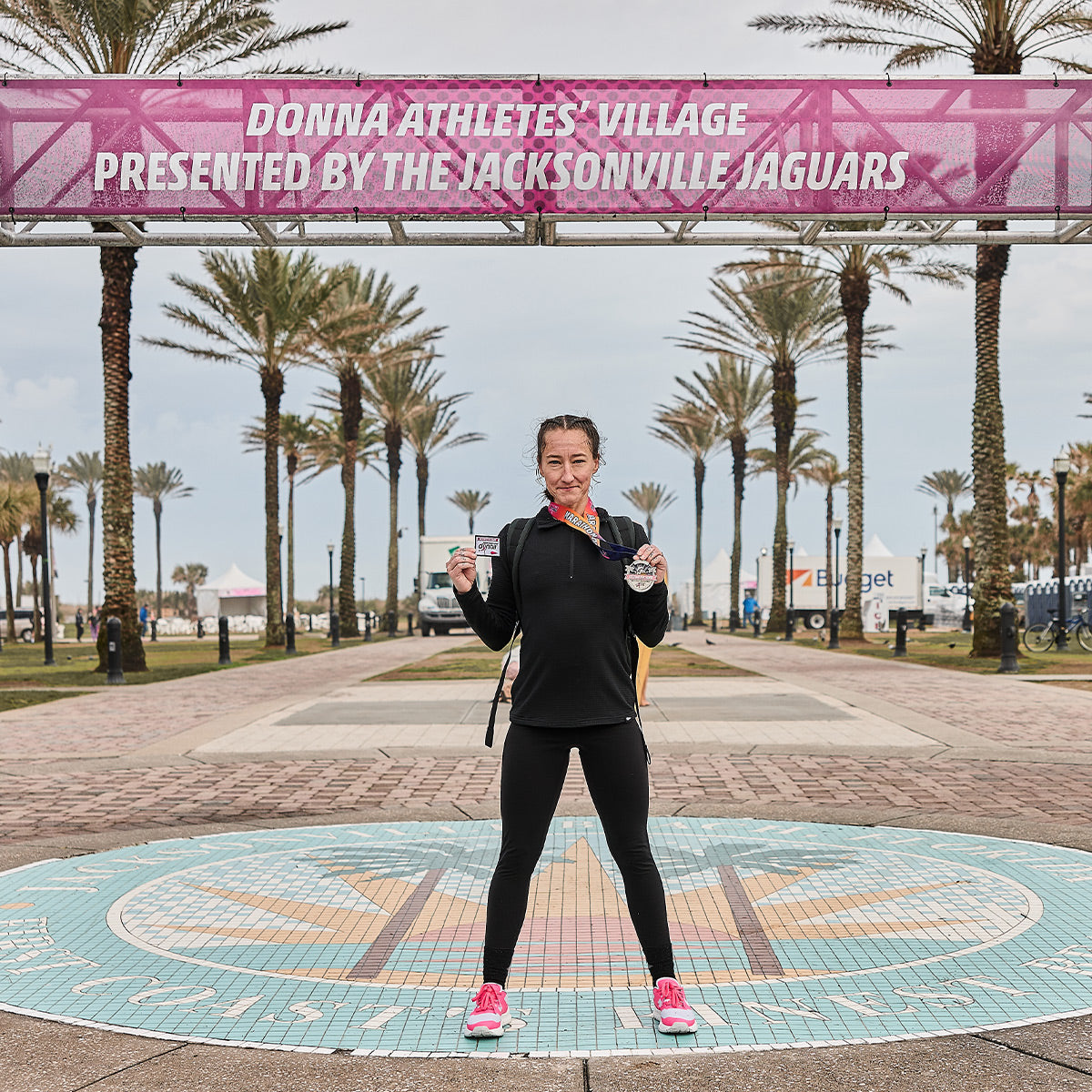 A runner stands proudly under a sign that reads Donna Athletes Village Presented by the Jacksonville Jaguars, holding a medal and water bottle. Women's Rough Runner - Hot Pink lightweight running shoes from GORUCK adorn their feet, while palm trees and a mosaic design on the ground create a vibrant setting.