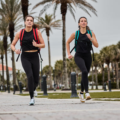 Two women jog along a palm tree-lined path, wearing black athletic outfits and colorful backpacks. Their Women's Rough Runner - Speed Tan shoes by GORUCK grip the ground effortlessly. The sky is cloudy, and the path is bordered by grass and lampposts, providing a perfect backdrop for these rough runners.