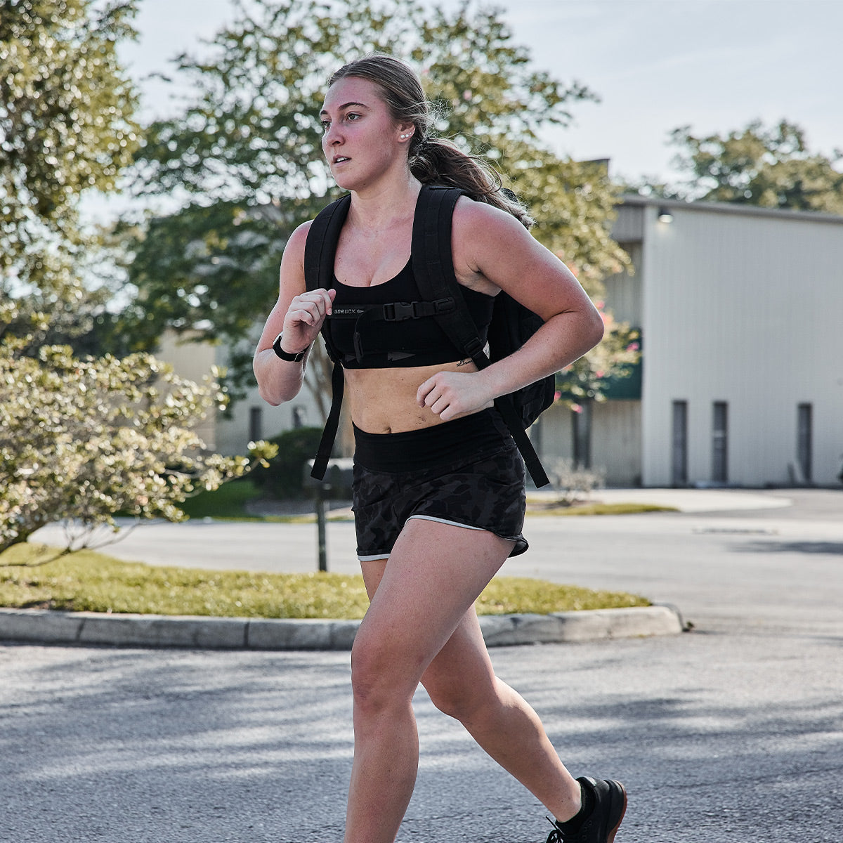 A woman jogs outdoors on a sunny day, wearing a fitness outfit featuring the Power Bra with Toughflex fabric from GORUCK, and carrying a weighted vest. Trees and a building stand in the background, enhancing her high-impact movements.