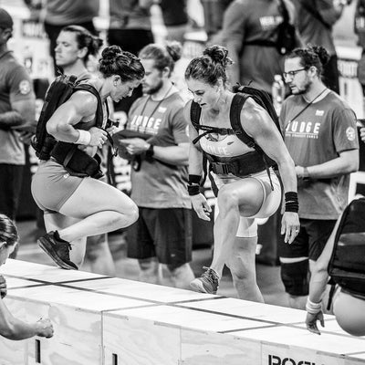 In a black and white photo, two athletes demonstrate agility and breathability by leaping over boxes during a fitness competition with Race Day Socks securely on their feet, while judges and spectators look on intently.