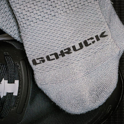 The gray Race Day Socks by GORUCK are shown on a black textured surface with part of a shoe visible. Designed for maximum breathability, these socks have reinforced high-friction areas to boost durability and performance.