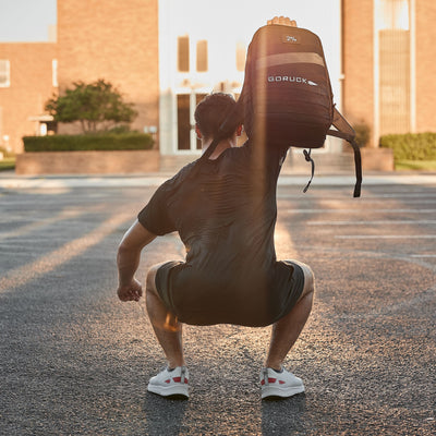 In a parking lot, sunlight casts long shadows as a person squats outdoors, holding a backpack overhead. Race Day Socks ensure optimal adventure performance with max breathability and reinforced high friction areas.