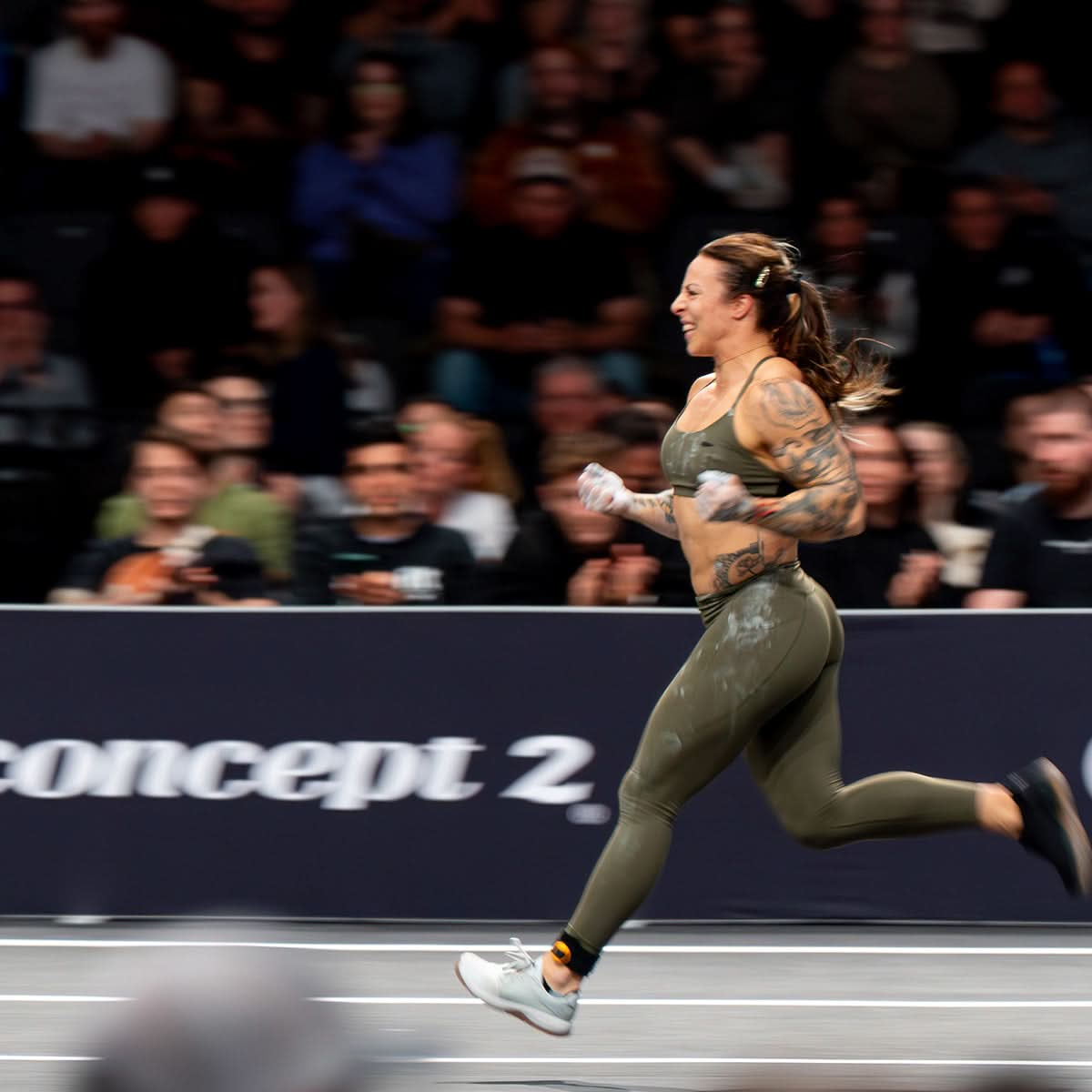 An athletic woman wearing green Race Day Socks with reinforced high friction areas runs energetically on an indoor track as spectators watch.