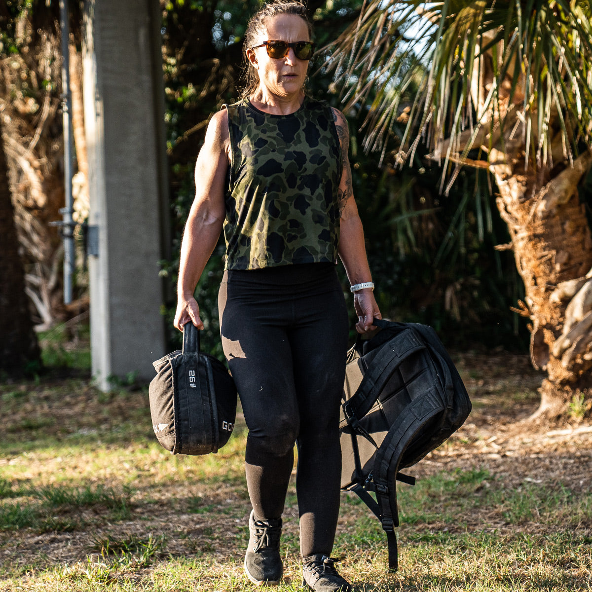 A person wearing sunglasses and a Women’s USA Performance Tank - ToughMesh from GORUCK carries a sandbag and backpack outdoors. They are walking on grass, with trees in the background, as the ToughMesh™ fabric dries lightning fast for ultimate comfort.