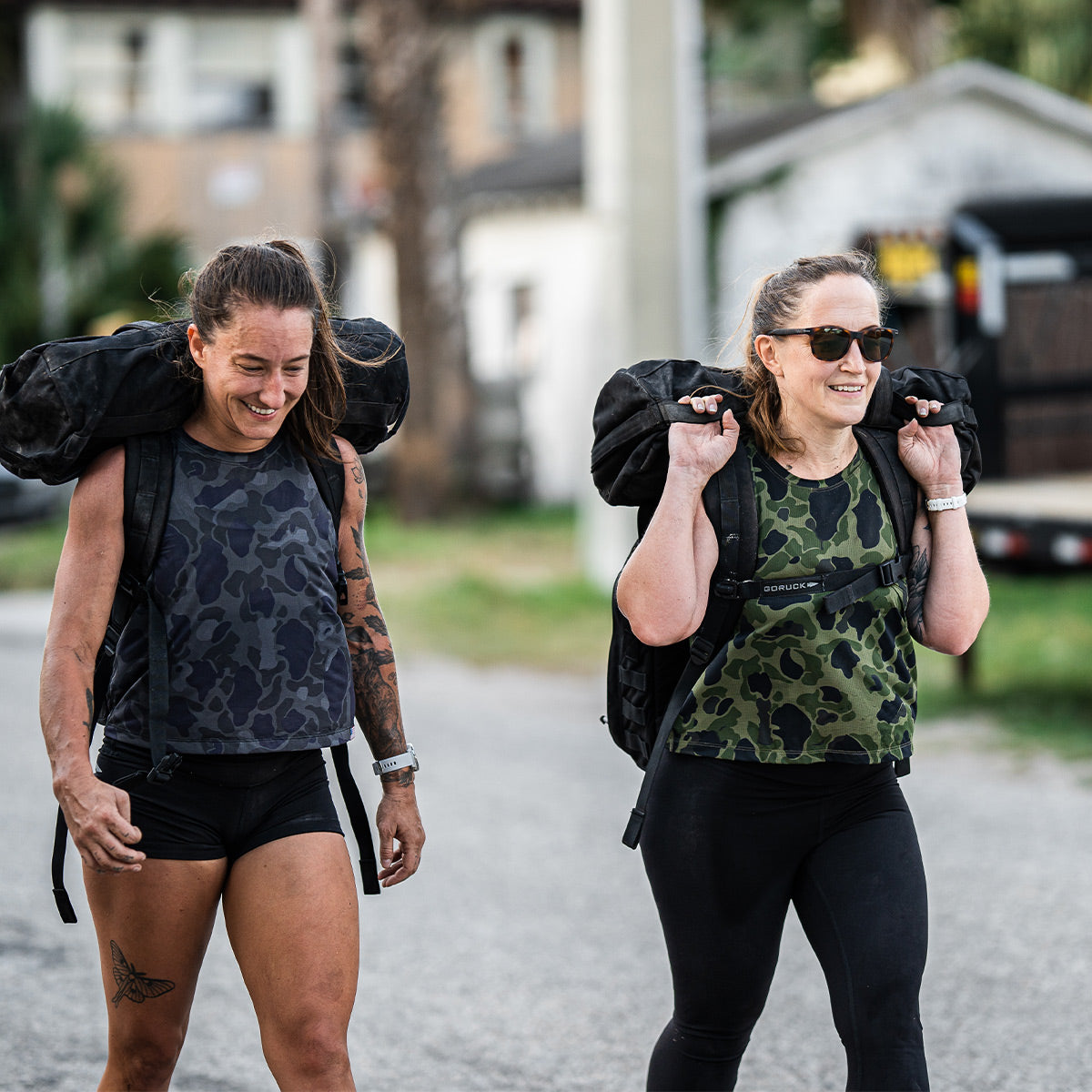 Two women wear the Women’s USA Performance Tank - ToughMesh by GORUCK in black and green camo, paired with shorts. They carry heavy backpacks while walking outdoors on a sunny day, smiling and chatting. One of them sports sunglasses. Buildings and greenery can be seen in the background, as their shirts dry lightning fast under the sun.