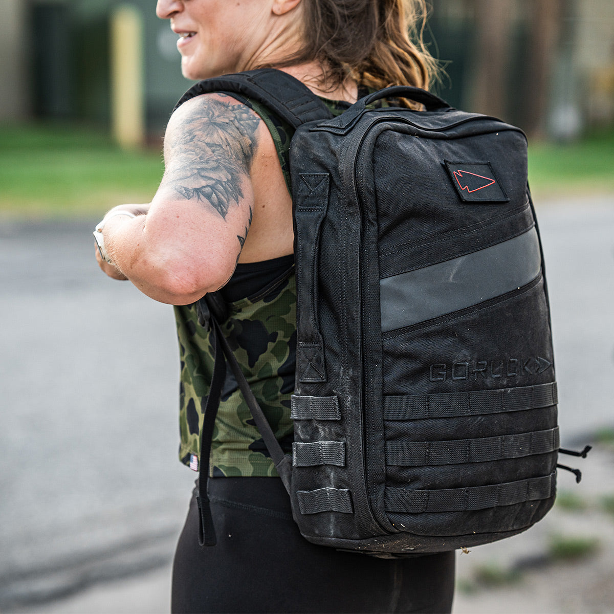 A person is wearing the Women’s USA Performance Tank by GORUCK, featuring a camo pattern and crafted with ToughMesh™ fabric that dries rapidly. They pair it with black leggings and carry a large backpack. With a tattoo on their arm, they stand outdoors on a paved surface.