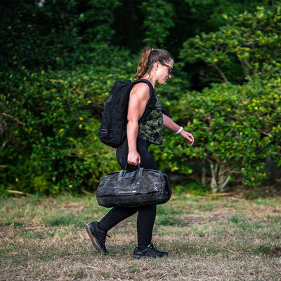 A woman in athletic gear, including a Women’s USA Performance Tank by GORUCK and a ToughMesh™ fabric backpack, as well as a black duffle bag, walks across a grassy area with lush green foliage in the background. Her featherweight training shirt ensures she moves effortlessly as it dries lightning fast.