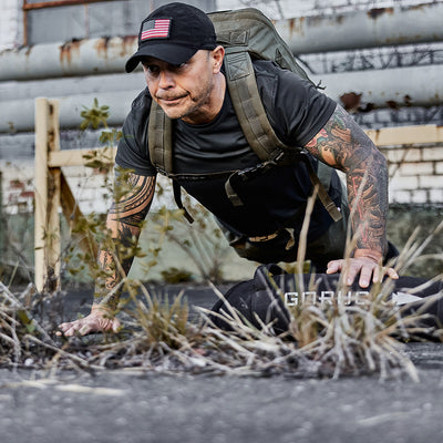 A man with a tattooed arm, wearing a black cap, performs push-ups outdoors with a GORUCK Rucker 4.0 backpack beside him. He is positioned on cracked pavement amid dry grass, and an industrial building stands prominently in the background.