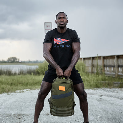 A man stands on a gravel path holding a green GORUCK Rucker 4.0 backpack. He is wearing a black T-shirt with a patterned America design, paired with black shorts. In the background, grassy fields stretch out alongside a fence under the cloudy sky, creating an ideal setting for rucking.